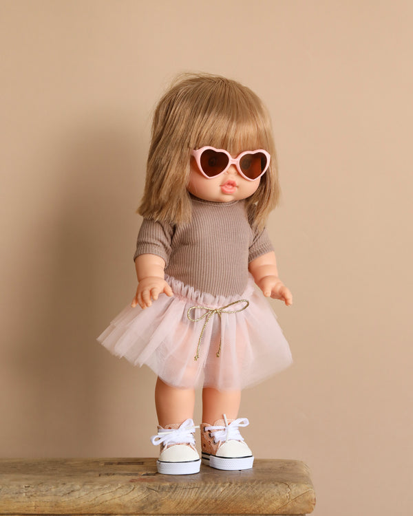 The Minikane Standing Doll, Zoelie in Pink Tutu, has long blonde hair and features pink heart-shaped sunglasses, a beige top, and white sneakers. Standing on a wooden surface against a beige backdrop, it emits a delightful natural vanilla scent. Ships in one week.