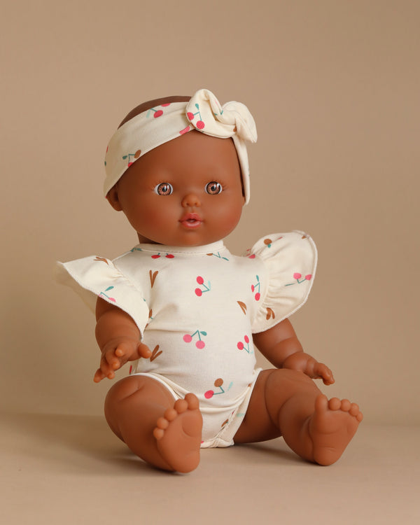 A Minikane Doll (13") - Léonie with Ondine Set in Cherries Outfit sits on a neutral background, featuring brown skin and brown eyes. Made of phthalate-free vinyl, the doll is dressed in a white outfit adorned with small, colorful cherry patterns and winged sleeves. Completing the look is a matching headband with a bow, making it perfect for imaginative playtime.