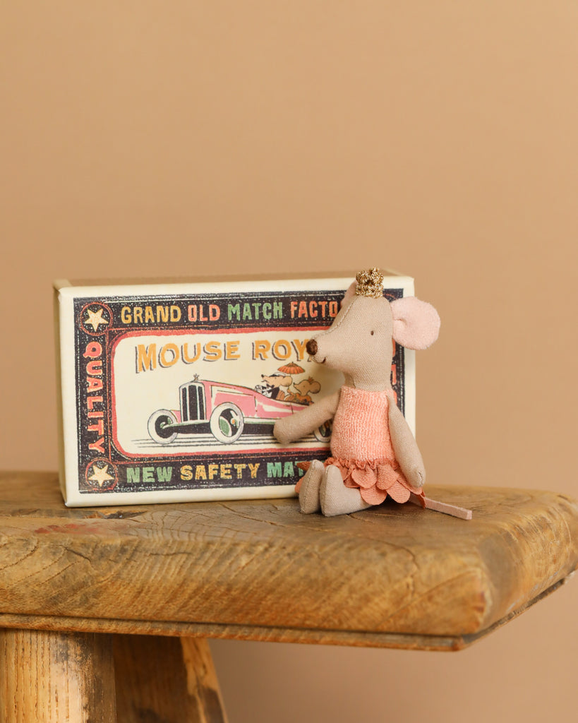 The Maileg Princess Mouse in Matchbox - Little Sister, dressed in a pink dress and crown, sits on a wooden surface exuding royal charm. Behind her is a matchbox bed with unique prints and a vintage car illustration labeled "Mouse Royal," adding whimsy to the scene.