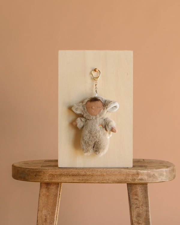 The Olli Ella Cozy Bag Charm - Lamby Pip, featuring sheep ears and a gray outfit, hangs on a wooden panel displayed atop a rustic stool against a plain beige background.