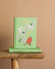 The mint green hardcover "The Wizard of Oz | Collector's Edition" showcases whimsical designs, including Emerald City and Yellow Brick Road motifs. It stands upright on a wooden stool against a beige background, with another edition lying flat below it.