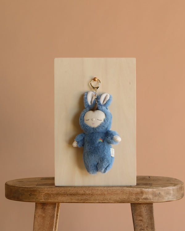 The Olli Ella Cozy Bag Charm - Bunny Twiggy, a plush blue bunny keychain with closed eyes and a rainbow on its chest, is displayed on a wooden board atop a rustic stool against a plain beige background.
