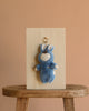 The Olli Ella Cozy Bag Charm - Bunny Twiggy, a plush blue bunny keychain with closed eyes and a rainbow on its chest, is displayed on a wooden board atop a rustic stool against a plain beige background.