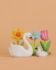 The Bumbu Handmade Wooden Swan Set, featuring large and small white swans, is displayed before three festive wooden flower decorations—yellow, blue, and pink—with green stems on a peach background.