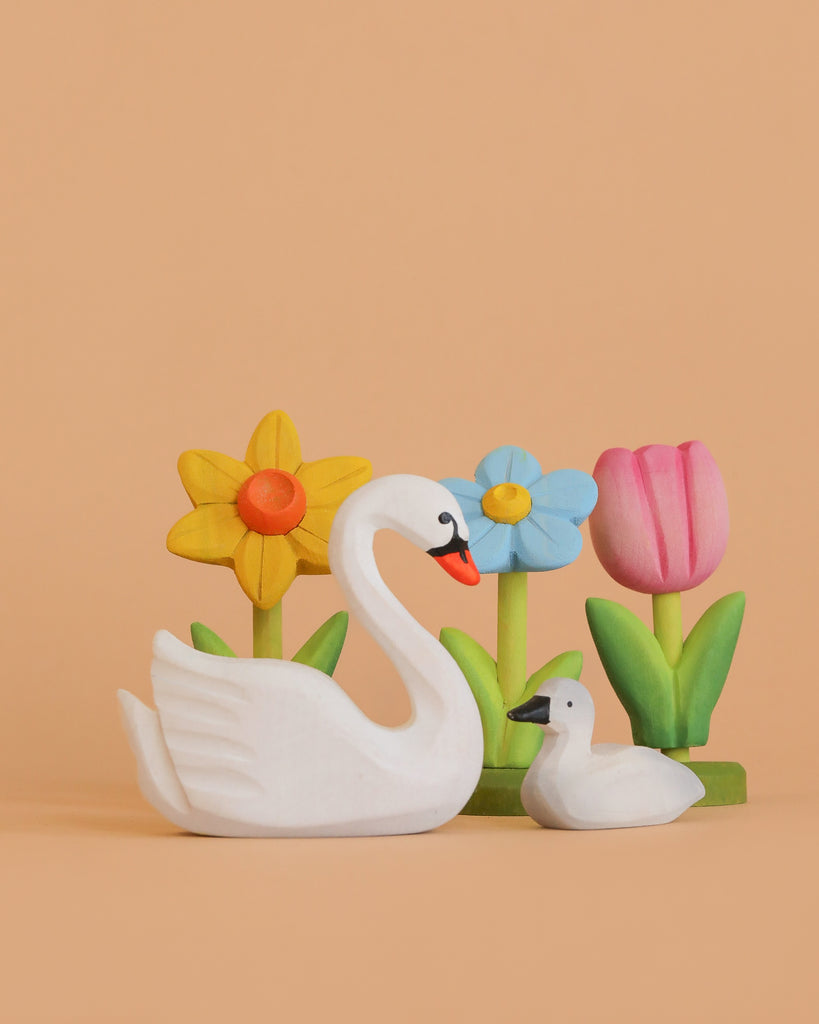 The Bumbu Handmade Wooden Swan Set, featuring large and small white swans, is displayed before three festive wooden flower decorations—yellow, blue, and pink—with green stems on a peach background.