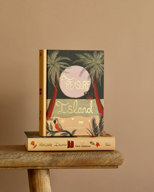 A hardcover collector's edition book titled "Treasure Island" by Stevenson is displayed upright on a wooden stool, along with a paperback version. Both feature palm tree and sailing ship illustrations, capturing the adventurous essence of this classic against a muted background.