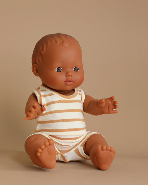 The Minikane Doll (13") - Léon with Marcel Set in Brown Sugar Stripes Outfit, made of phthalate-free vinyl, features a brown-skinned baby doll with short hair seated on a neutral-colored surface. Dressed in a light-colored sleeveless outfit with beige striped patterns, it extends both arms forward, ready for imaginative playtime.