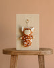 The Olli Ella Cozy Bag Charm - Fawny Lala features a cute animal with closed eyes, orange fur, white spots, and ears. Displayed on a light wooden board atop a stool against a beige background.