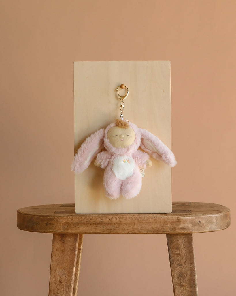 The Olli Ella Cozy Bag Charm - Bunny Flopsy, a soft pink bunny with closed eyes, hangs on a wooden board placed atop a wooden stool against a light brown backdrop.