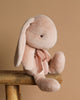 The Maileg Plush Bunny - Medium Light Powder, a soft beige bunny with unique fabric, floppy ears, and a neck ribbon, sits on a wooden stool against a light brown background.