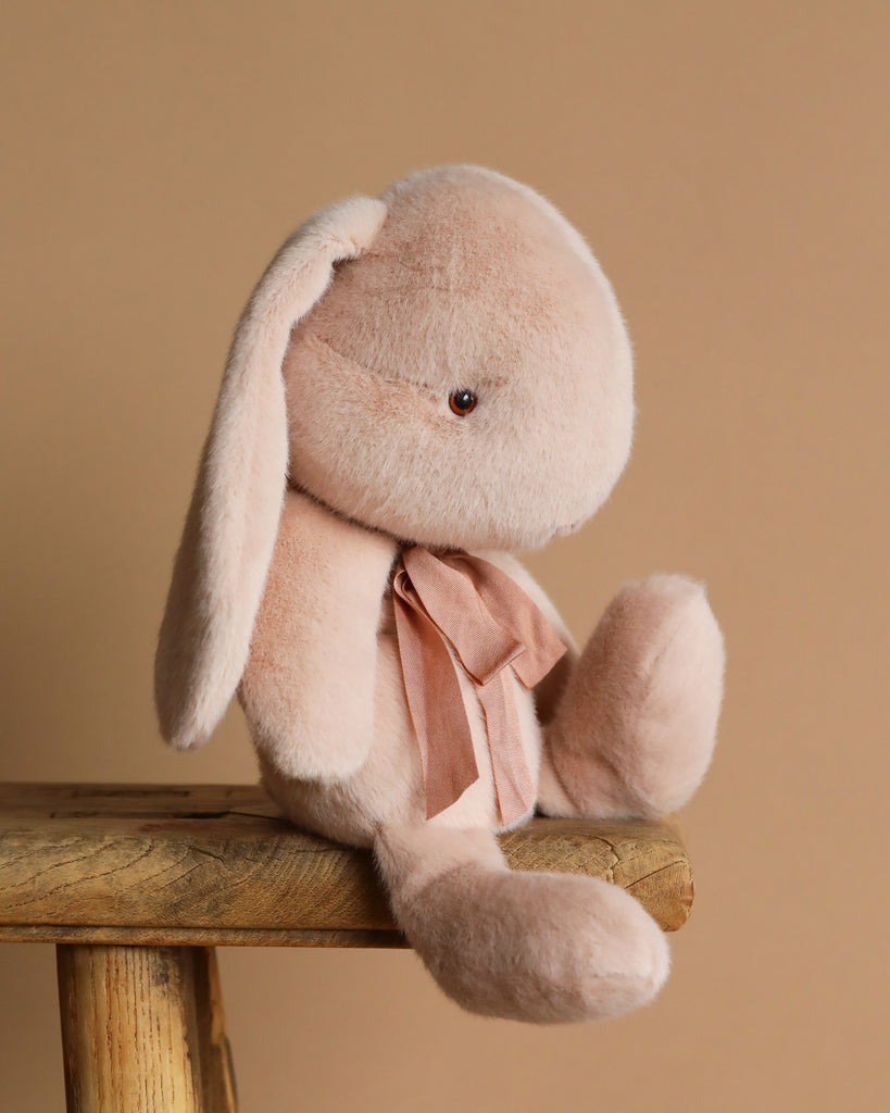 The Maileg Plush Bunny - Medium Light Powder, a soft beige bunny with unique fabric, floppy ears, and a neck ribbon, sits on a wooden stool against a light brown background.