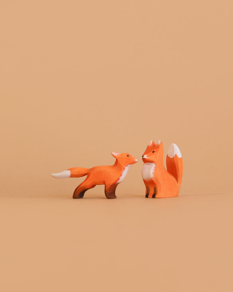 The Bumbu Handmade Wooden Fox Set features two small, orange wooden fox figurines with white accents—one sitting upright, the other standing with its tail extended—perfect for eco-friendly Montessori imaginative play.