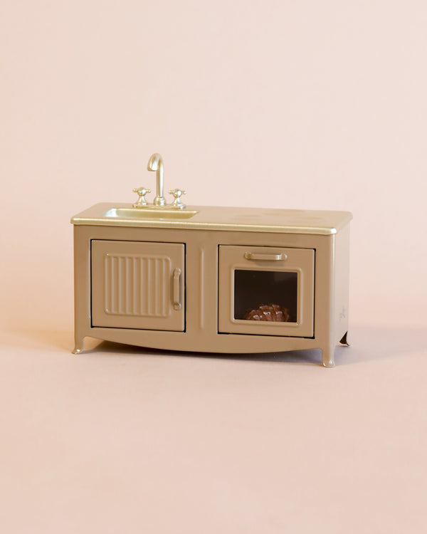 A Maileg Kitchen - Light Brown model with a metal sink, faucet, and a cabinet door with a clear window revealing dishes inside, set against a pale pink background.