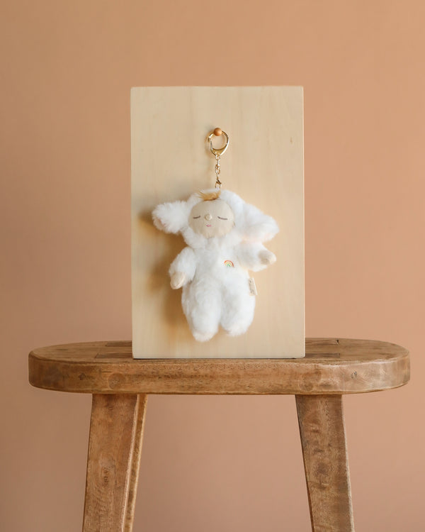 The Olli Ella Cozy Bag Charm - Lamby Pookie, a fluffy white plush keychain with floppy ears and a rainbow patch, hangs on a wooden board set on a stool against a light brown background.