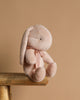 The Maileg Plush Bunny - Small Light Powder, made from recycled polyester with long ears and light beige fur, adorned with a pink ribbon around its neck, sits on the edge of a wooden stool against a plain beige background.