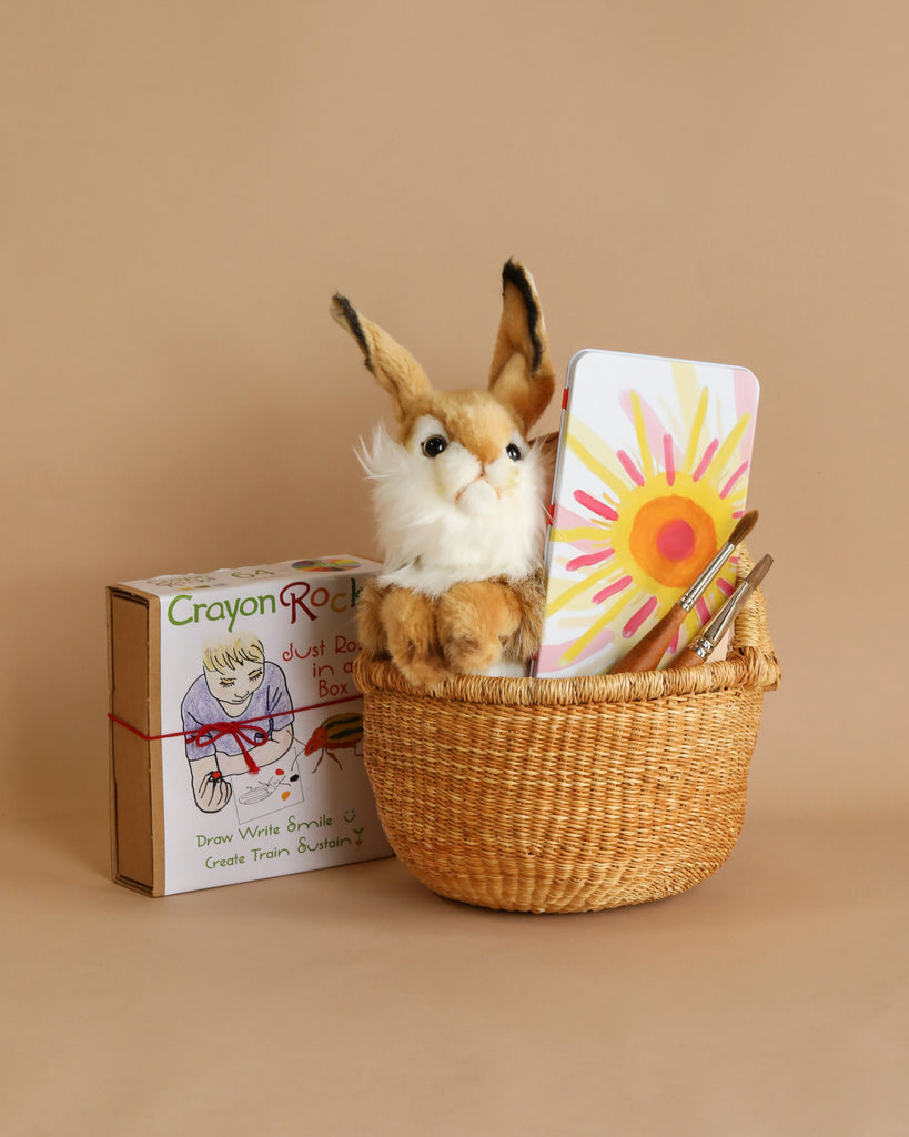 A plush rabbit toy sits in an Easter Basket Set, holding a smartphone displaying a child's drawing next to a box of Crayon Rocks and markers with creative designs on it. A beige background comple