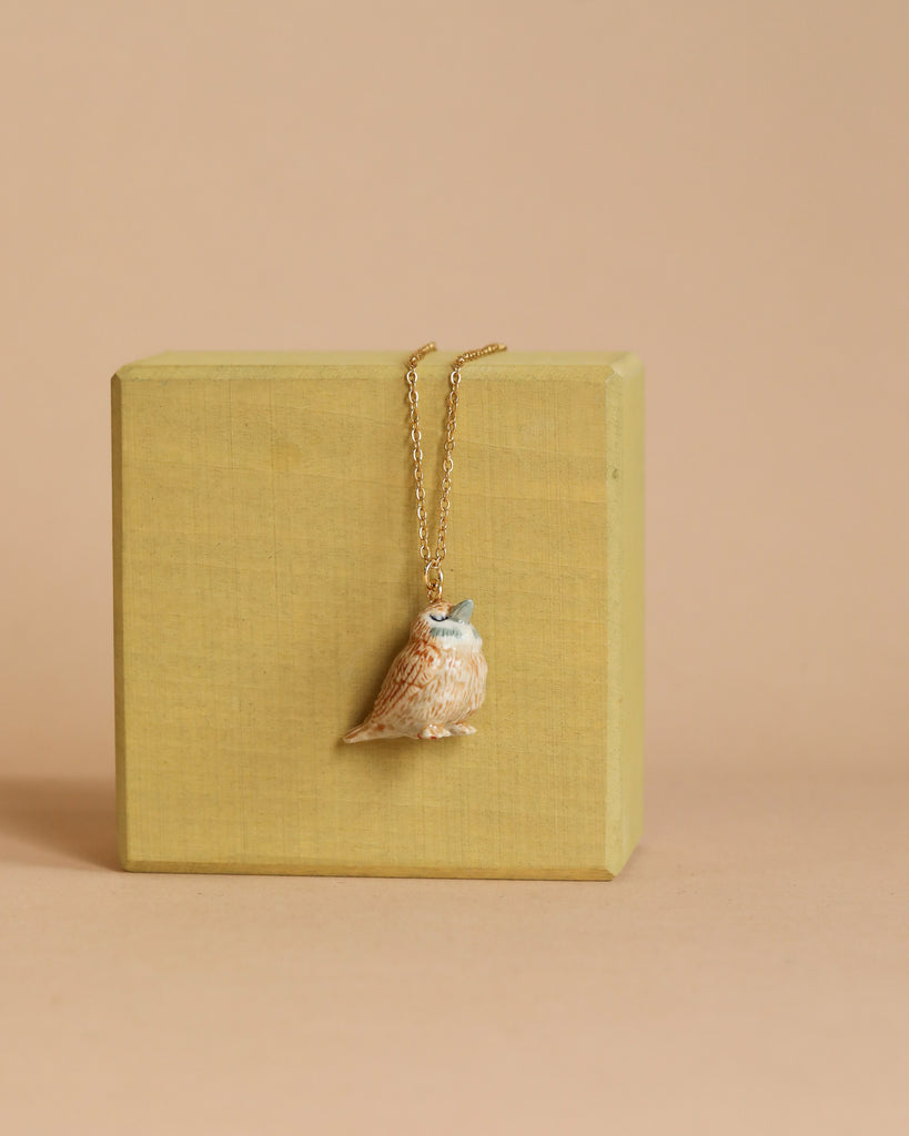 A wooden cube serves as a display stand for the limited-edition Sparrow Necklace, featuring an heirloom-quality pendant shaped like a small owl. The owl pendant boasts a detailed design and is primarily light brown with subtle blue accents around its eyes and beak. The background is plain beige.