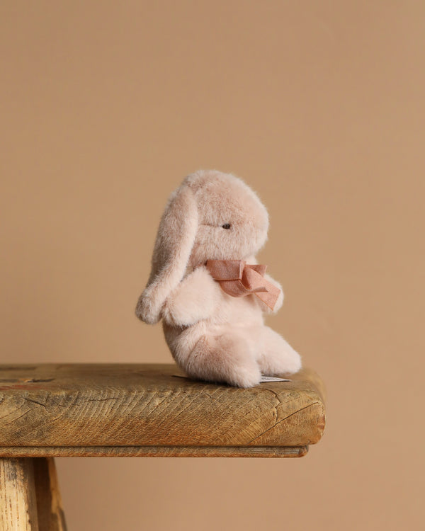 The Maileg Plush Bunny - Mini Light Powder, with its soft fabric, floppy ears, and pink bow, adorns the edge of a wooden stool against a solid beige backdrop, creating a warm and cozy atmosphere.