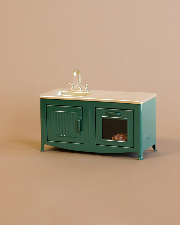 The Maileg Kitchen - Dark Green is a charming miniature kitchen unit with a white top, sink, and faucet. Below are two detailed doors: one ribbed, the other with a window showing food inside, perfect for a mouse family against a plain beige background.