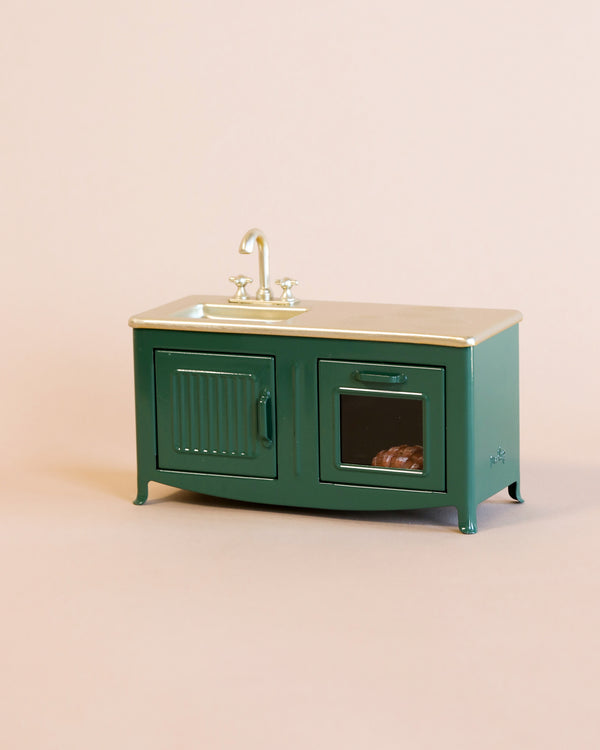 A miniature Maileg Kitchen - Dark Green with a sink and faucet on top, featuring one visible open cabinet door displaying a freshly baked pie inside. The background is plain and light pink.