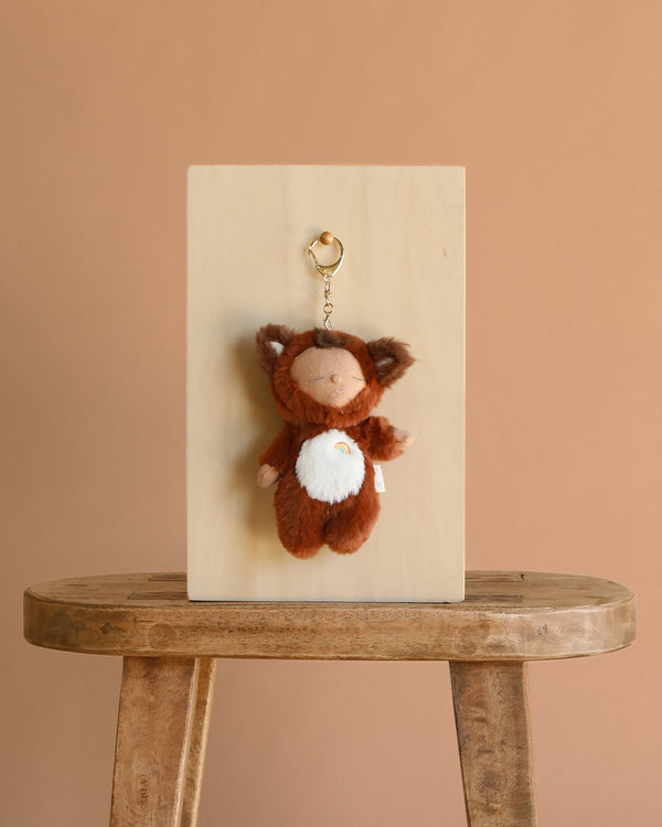 The Olli Ella Cozy Bag Charm - Fox Finnley, a plush brown fox with a white belly, hangs on a wooden board atop a rustic stool against a plain beige background.