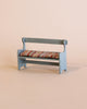 A small Maileg Christmas Mini Bench painted light blue with curved armrests and a multi-colored striped cushion on the seat gives off a vintage look, set against a plain beige background.