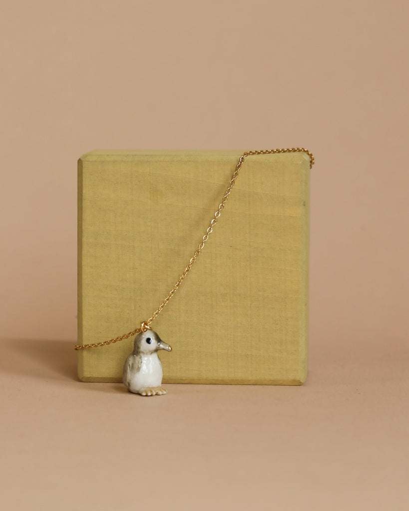 A Tiny Penguin Necklace with a gold-tipped beak hangs gracefully on a delicate gold chain, draped over a plain wooden block set against a beige background. The penguin pendant is white with subtle detailing, adding a whimsical touch to the handmade jewelry presentation.