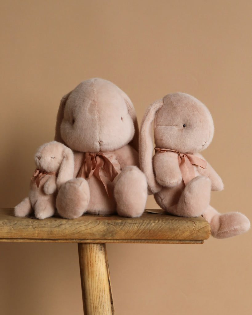 Three Maileg Plush Bunnies in light powder, crafted from recycled polyester, sit on a wooden stool against a brown background. Featuring long ears and pink bows, they vary in size with the smallest on the left and largest on the right.