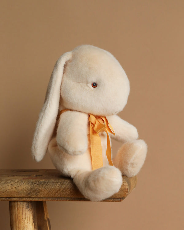 A Maileg Plush Bunny in medium cream, crafted from soft plush and recycled polyester, is seated with long ears and a small orange ribbon on a wooden stool against a plain brown backdrop.