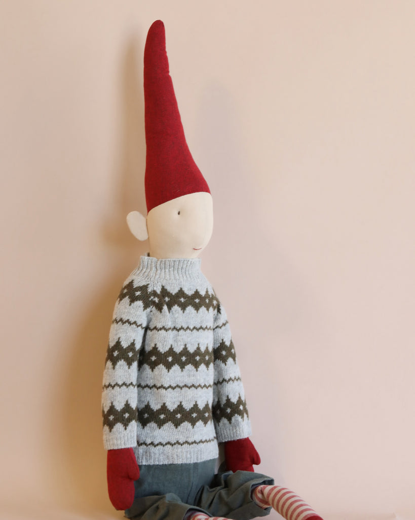A Maileg Christmas Mega Pixy - (Size 6) with a long red hat, wearing a gray sweater with a white and black geometric pattern, against a light pink background, designed as an exclusive Christmas decoration.