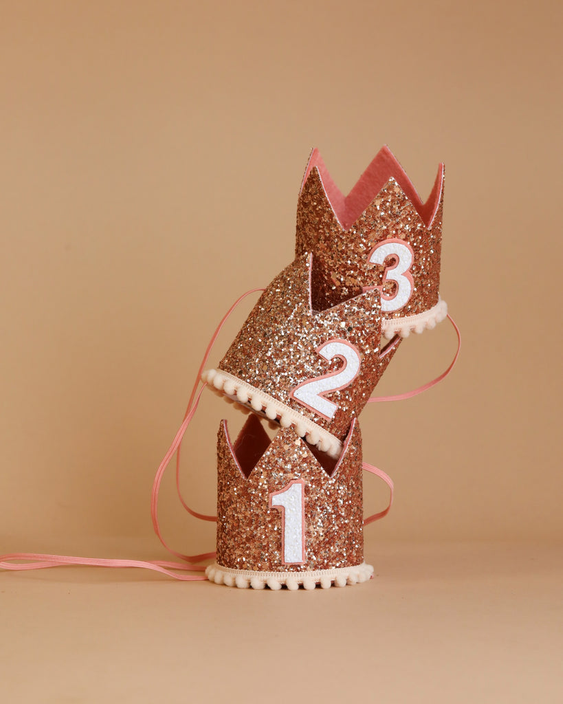 Three small Rose Gold Glitter Crowns, each featuring a number (1, 2, and 3) on the front, are stacked together against a beige background. Perfect for birthday celebrations, these crowns are adorned with white trimming at the base and have pink ribbons attached.