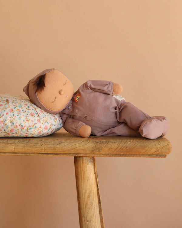 The Olli Ella Dozy Dinkum - Pip, a soft cotton doll in mauve, rests on a floral pillow atop a wooden bench against a beige background—perfect as baby's first toy.