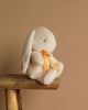 The Maileg Plush Bunny - Small Cream, with long ears and an orange ribbon around its neck, sits on a rustic wooden bench. Made from recycled polyester, it evokes a warm, cozy ambiance against the plain beige background.