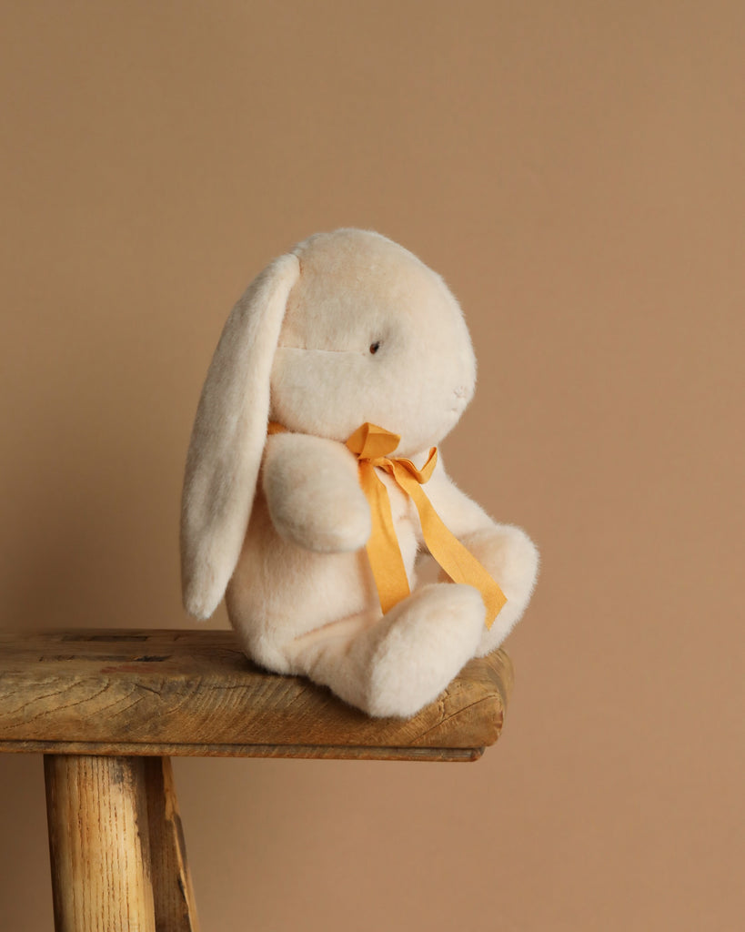 The Maileg Plush Bunny - Small Cream, with long ears and an orange ribbon around its neck, sits on a rustic wooden bench. Made from recycled polyester, it evokes a warm, cozy ambiance against the plain beige background.