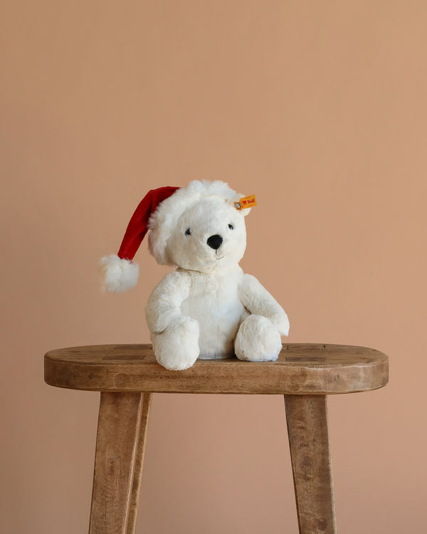 The Steiff Cozy Christmas Rico Polar Bear, wearing a red Santa hat, sits on a wooden stool against a beige background, adding festive charm to the scene.