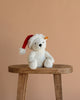 The Steiff Cozy Christmas Rico Polar Bear, wearing a red Santa hat, sits on a wooden stool against a beige background, adding festive charm to the scene.