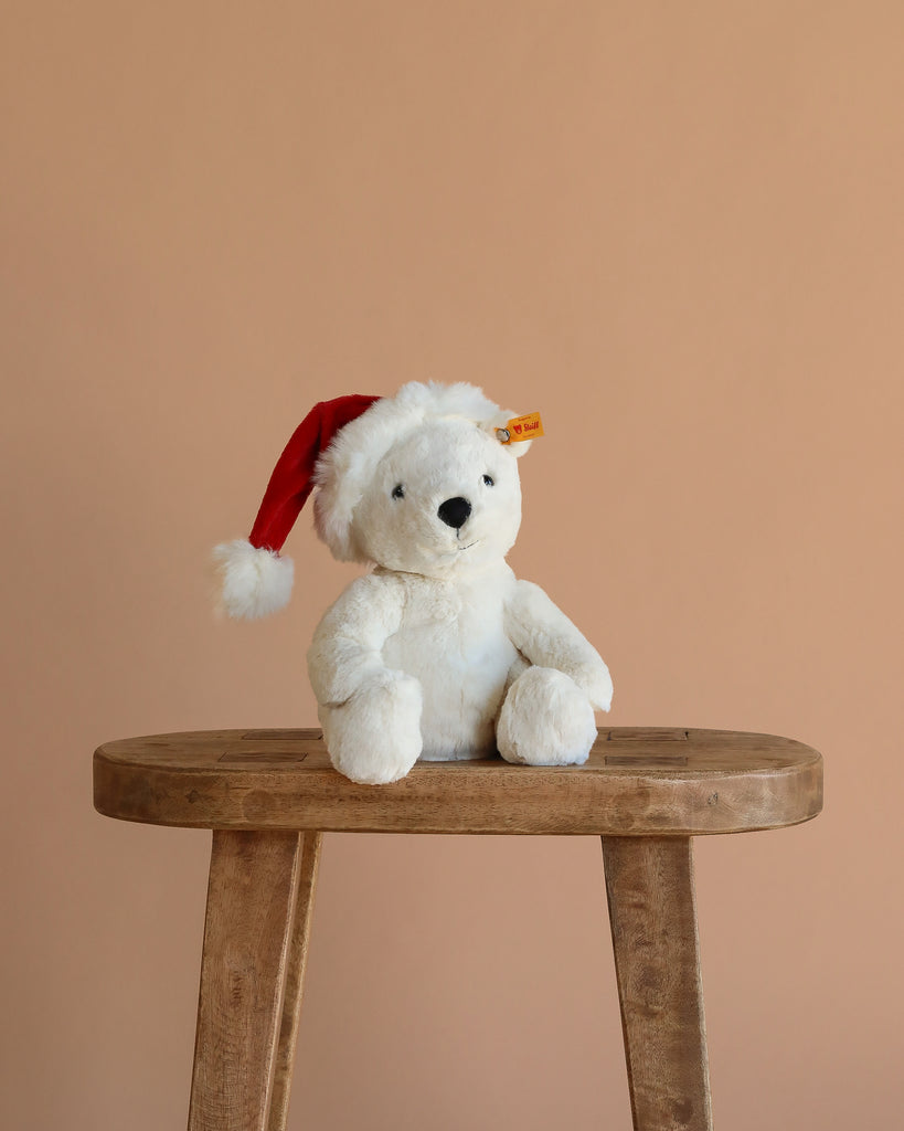 The Steiff Cozy Christmas Rico Polar Bear, wearing a red Santa hat, sits on a wooden stool against a beige background, adding festive charm to the scene.