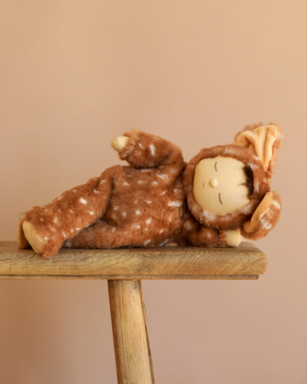The Olli Ella Cozy Dinkum - Fawny Twiggy, a plush doll in a brown deer-themed outfit with white spots, rests on its side atop rustic wood with a beige backdrop—a charming collectible and perfect holiday gift.
