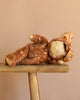 The Olli Ella Cozy Dinkum - Fawny Twiggy, a plush doll in a brown deer-themed outfit with white spots, rests on its side atop rustic wood with a beige backdrop—a charming collectible and perfect holiday gift.