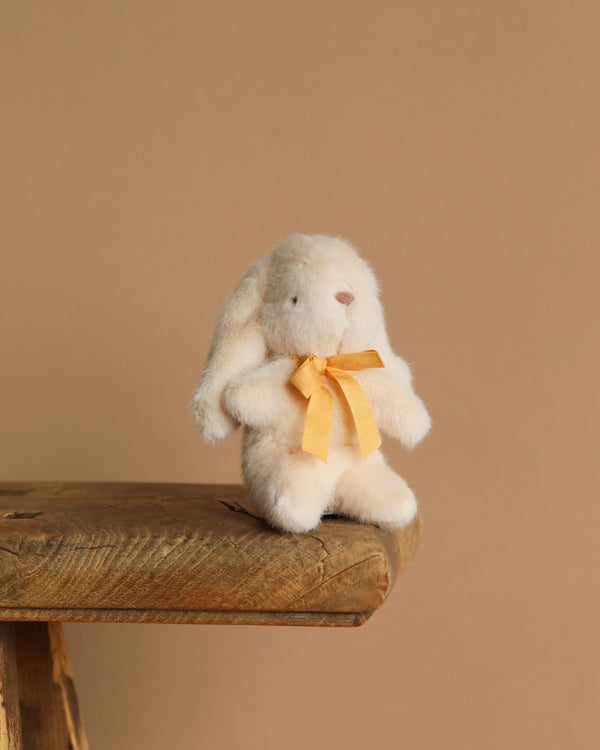 The Maileg Plush Bunny - Mini Cream, featuring a yellow ribbon around its neck, sits on the edge of a rustic wooden bench, crafted from unique plush fabric against a plain beige background.