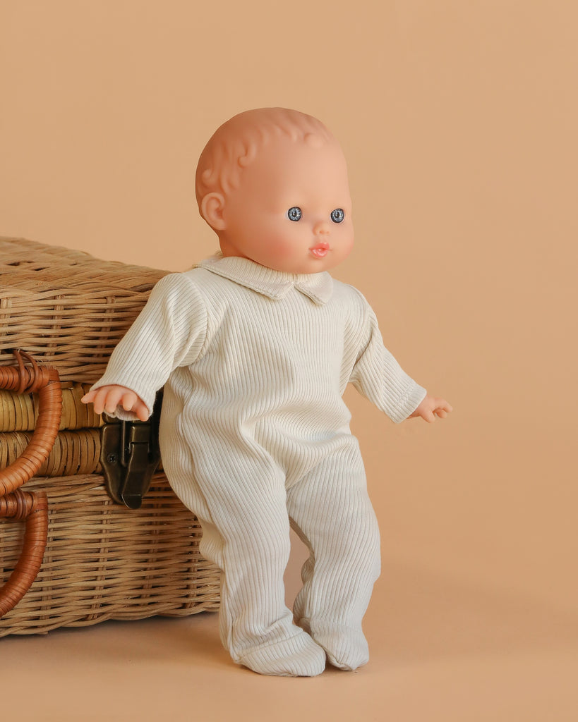 The Minikane Soft Body Doll (11") - Claire with Clothing stands upright in a white ribbed outfit next to a wicker basket against a beige background.