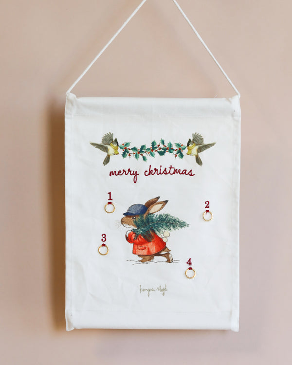 A Christmas Advent Calendar hanging on a wall, featuring an illustrated robin holding an apple, with numbered pockets and small gold rings. Floral and bird embellishments with "merry christmas" text at the bottom.