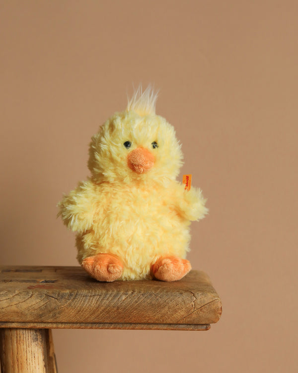 The Pipsy Chick Stuffed Plush Spring Animal, resembling a cheerful yellow ball of fur with orange feet, a beak, and a tuft of white hair, sits on a wooden surface against a beige background. Perfect for the "Friends Under $50" collection as an Easter Basket addition.