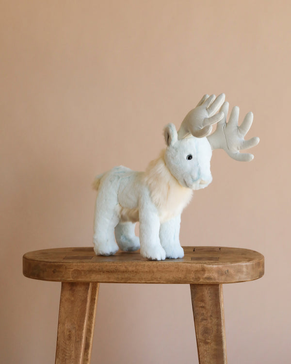 The Steiff Harry Potter "Patronus Stag" 7" plush, a light blue and white stuffed animal with large antlers, stands on a wooden stool against a plain beige background.