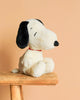 A Steiff Peanuts Snoopy plush with black ears and a red collar sits on a wooden stool against a plain peach-colored background.