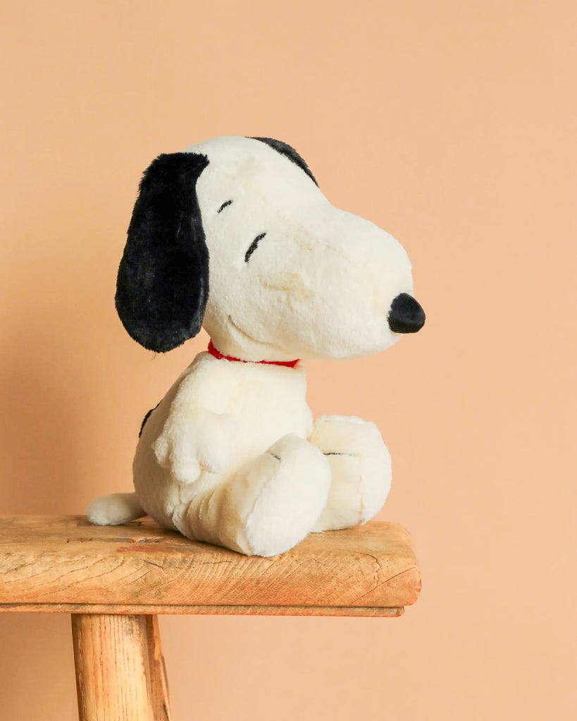 A Steiff Peanuts Snoopy plush with black ears and a red collar sits on a wooden stool against a plain peach-colored background.
