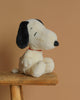 The Peanuts Snoopy Plush Toy, featuring a white body, black ears, and a red collar, is positioned on a wooden stool in front of a beige background.