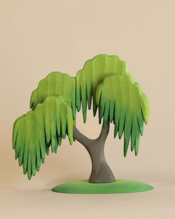 A small, stylized wooden tree with a curved trunk and drooping, layered green foliage stands on a flat green base against a plain beige background. This Large Wooden Willow Tree is painted with non-toxic paint, featuring a minimalist design that appears to be handcrafted.