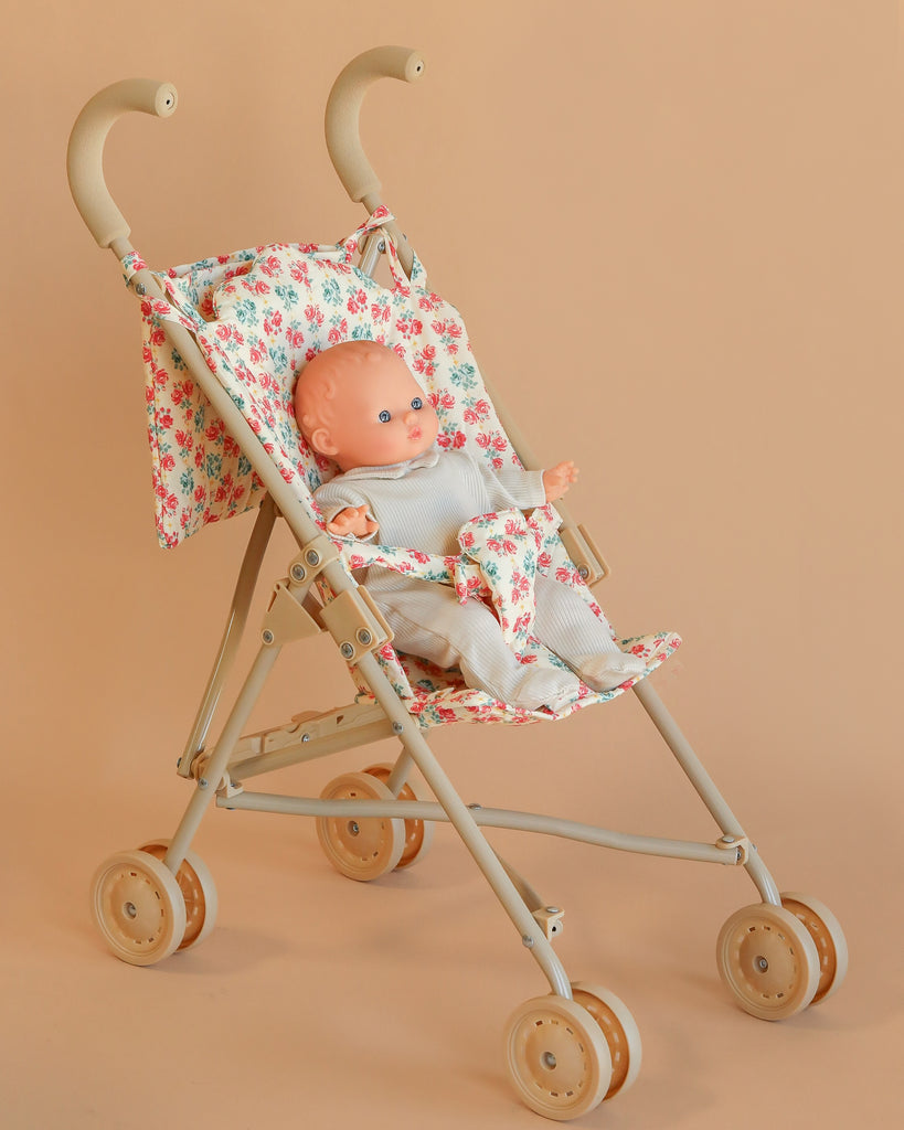The Minikane Soft Body Doll (11") - Claire with Clothing and Stroller is displayed in a beige toy stroller with floral fabric, set against a peach-colored background.