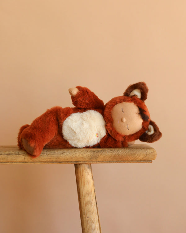 A plush toy with a sleeping face, dressed in a fuzzy fox costume, lies on its back atop a wooden bench against a plain beige background—meet the snuggly Olli Ella Cozy Dinkum Doll - Fox.